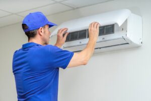 A man in a blue polo shirt and blue hat reaches up to adjust an air conditioning system attached to the top of a wall.